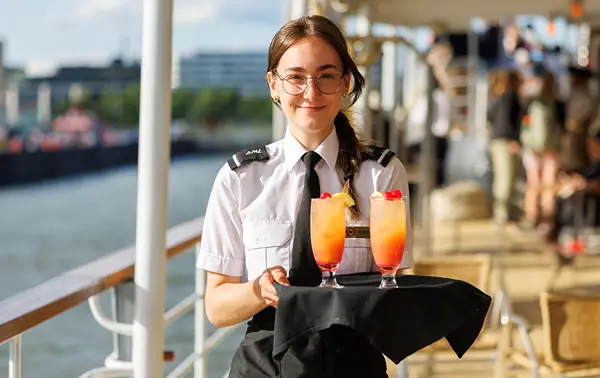 A smiling woman wearing round sunglasses and a striped shirt, holding a green beer bottle, with a river and blurred buildings in the background.