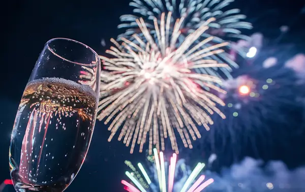 A champagne glass raised in the foreground while multicolored fireworks light up the night sky in the background.