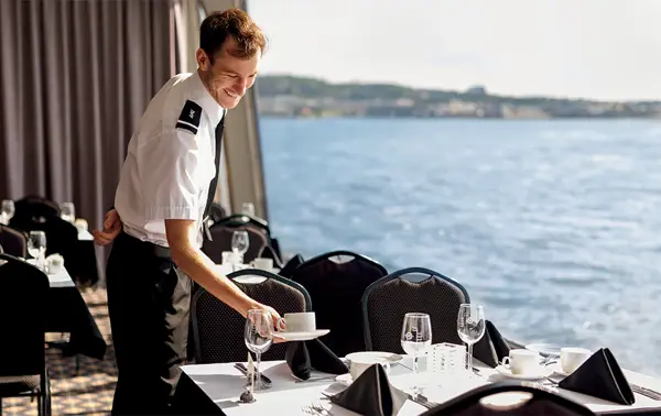 A uniformed waiter sets an elegant table with white tablecloths and black napkins in a dining room with a panoramic view of the water in the background.