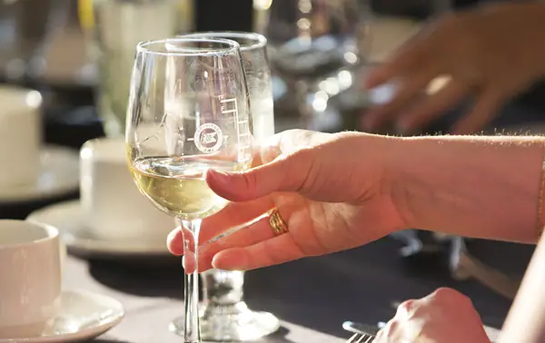 A hand holds a glass of white wine illuminated by sunlight, with an elegant table in the background featuring cups and glasses.