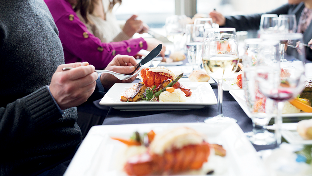 Gros plan sur des gens qui dînent avec une assiette de homard au premier plan accompagnée de verres de vin et d'autres plats autour. 