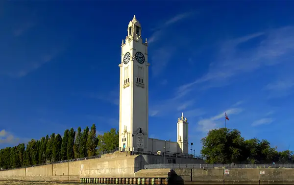 A tall white clock tower located on a quay, surrounded by trees and under a clear blue sky. A small structure and a flag are visible nearby.