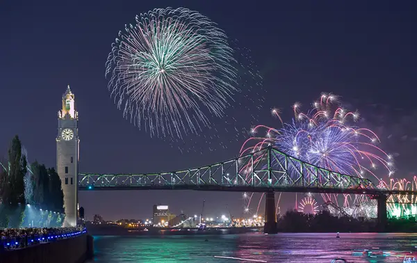 A spectacular nighttime view of fireworks lighting up the sky above an iconic bridge and a clock tower, with sparkling city lights and a calm river in the foreground.