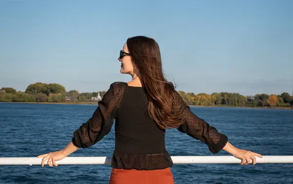 A woman viewed from behind, wearing a black blouse and sunglasses, leaning on a railing, admiring a river with trees and a clear sky in the background.
