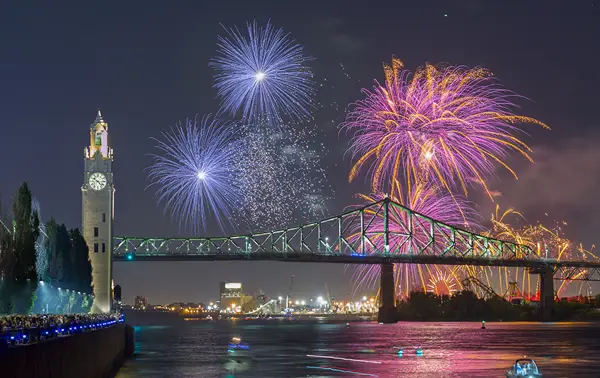 Colorful fireworks burst in the night sky near the Jacques Cartier Bridge and the Clock Tower, with reflections on the river and a crowd watching from the quay.