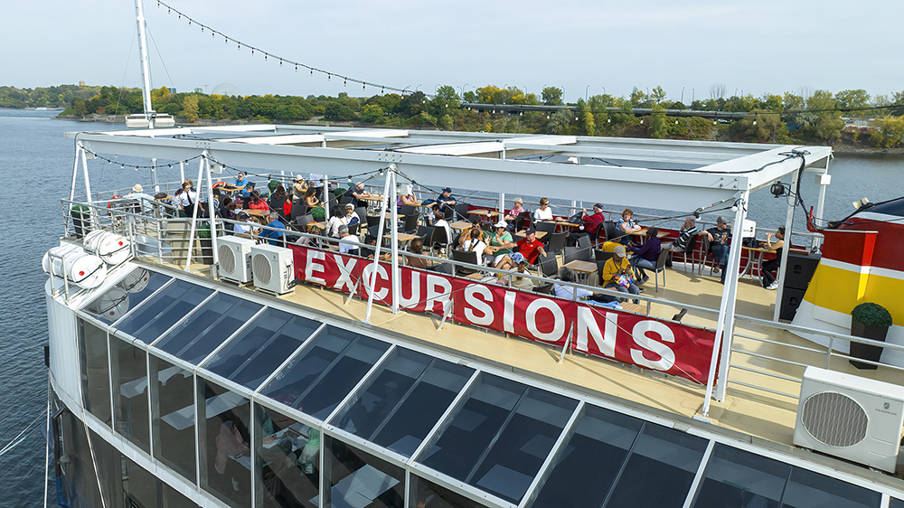 Outdoor terrace on the upper deck of the AML Cavalier Maxim ship, with customers seated at tables enjoying the view of the St. Lawrence River and Île Sainte-Hélène in the background.