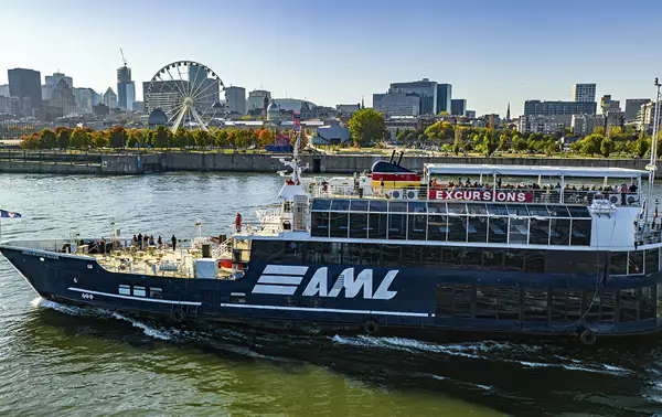 A large black cruise ship with "AML" written on it, sailing on a river. In the background, a Ferris wheel, autumn trees, and urban buildings under a clear blue sky.
