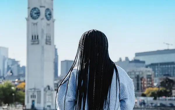 A woman viewed from behind with long black braids, looking at a tall white clock tower and a cityscape with modern buildings under a clear blue sky.