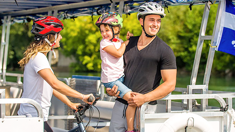A couple and a child enjoying a ride on a river shuttle on the St. Lawrence River in Montreal. The woman is holding a bicycle and wearing a helmet, while the man is carrying the child in his arms.
