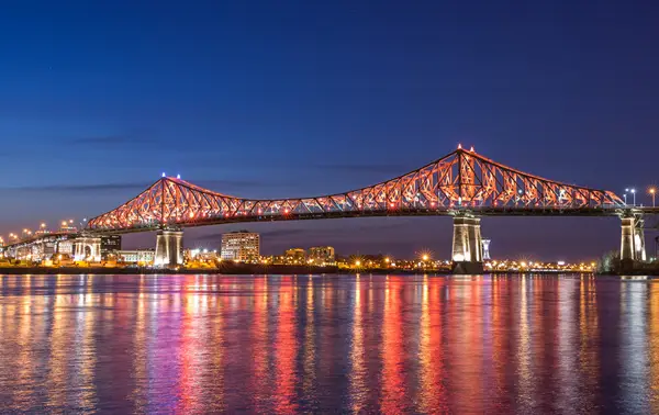 The Jacques-Cartier Bridge lit up in red and white, reflecting on a calm river at night, with city lights and a deep blue sky in the background.