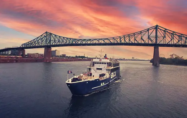A blue cruise ship labeled "AML" sails on a river at sunset, with the Jacques-Cartier Bridge in the background under a stunning sky with pink and orange hues.
