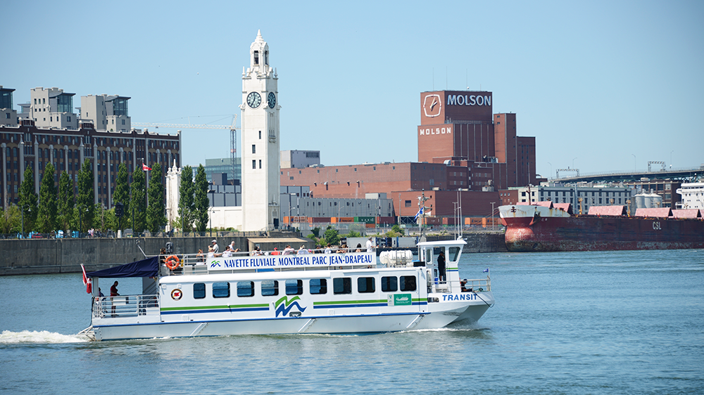 A river shuttle sailing on the St. Lawrence River, with the river in the foreground and the Clock Tower and the Molson Brewery building in the background.