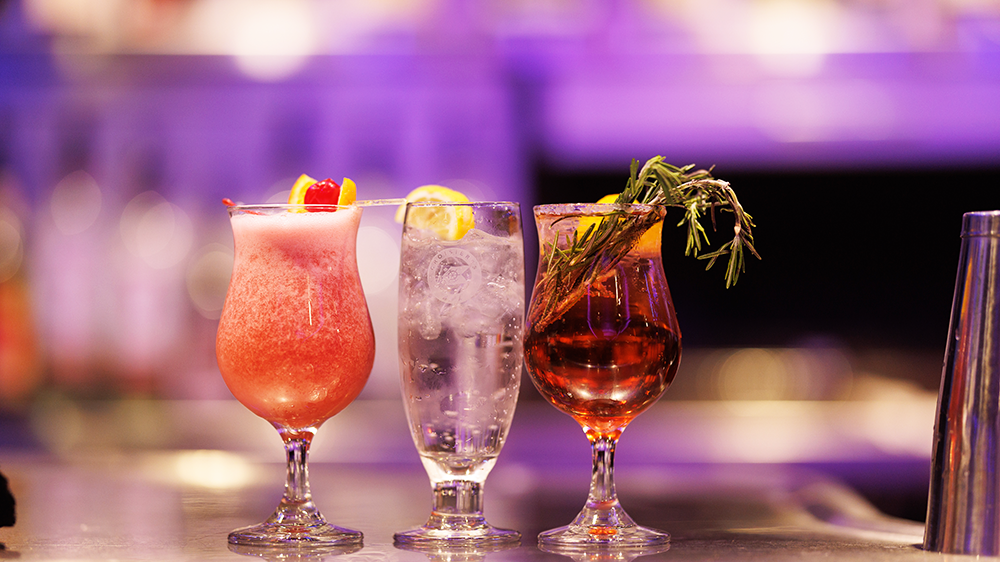  Three cocktails of various colors and garnishes on a bar counter, with a slightly blurred background highlighting the vibrant drinks.