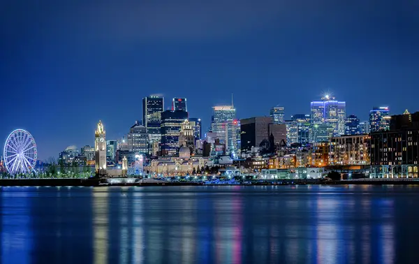 Nighttime view of a city illuminated with skyscrapers, a lit-up Ferris wheel, and reflections on the calm water in the foreground.