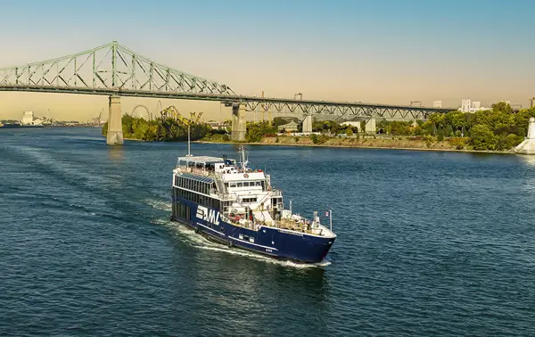 A cruise ship sails on a river on a sunny day, with the Jacques-Cartier Bridge in the background and trees lining the shore.