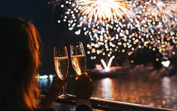 Two people toast with champagne glasses on a balcony, enjoying a spectacular fireworks display lighting up the night sky and reflecting on the water.