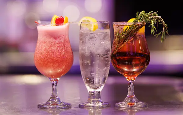Three elegant cocktails lined up on a counter, featuring a pink cocktail garnished with a cherry, a clear drink with a lemon slice, and an amber cocktail decorated with rosemary.