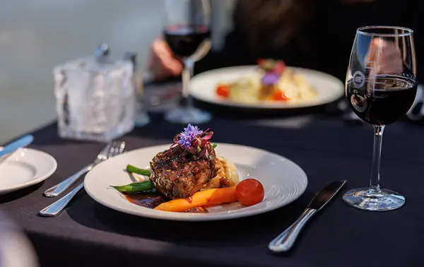 A beautifully plated dish with a piece of meat garnished with vegetables and edible flowers, accompanied by glasses of red wine on a table with a dark tablecloth.