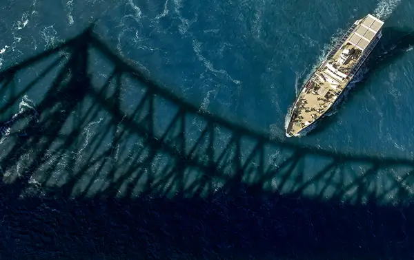Aerial view of a boat sailing on a blue river, with the imposing shadow of a bridge cast on the water.