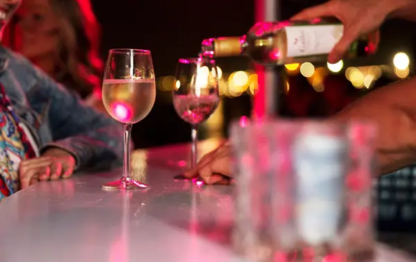 A server pours white wine into glasses at a bar under soft pink lighting, with smiling customers in the background.
