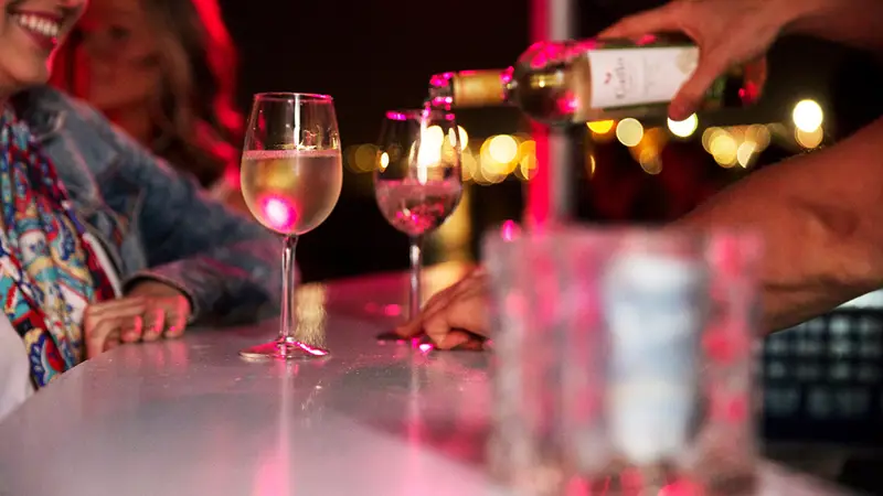 A server pours white wine into glasses at a bar under soft pink lighting, with smiling customers in the background.