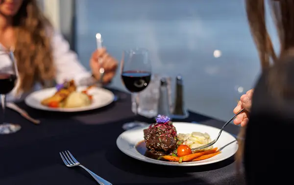Two people enjoying a gourmet meal on an elegant table with a black tablecloth, including red wine and beautifully plated dishes, with a view of the water in the background.