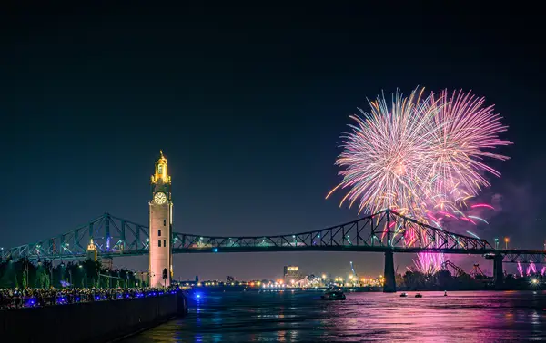Colorful fireworks burst in the night sky near the Jacques Cartier Bridge and the Clock Tower, with reflections on the river and a crowd watching from the quay.