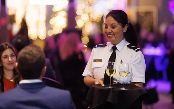 A smiling waitress in uniform serves two glasses of white wine to guests in an elegant setting with dim lights and a festive atmosphere.