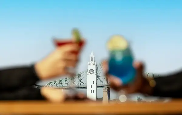Two people toasting with colorful cocktails (one red and one blue) against a blurred background of a white clock tower and a bridge.