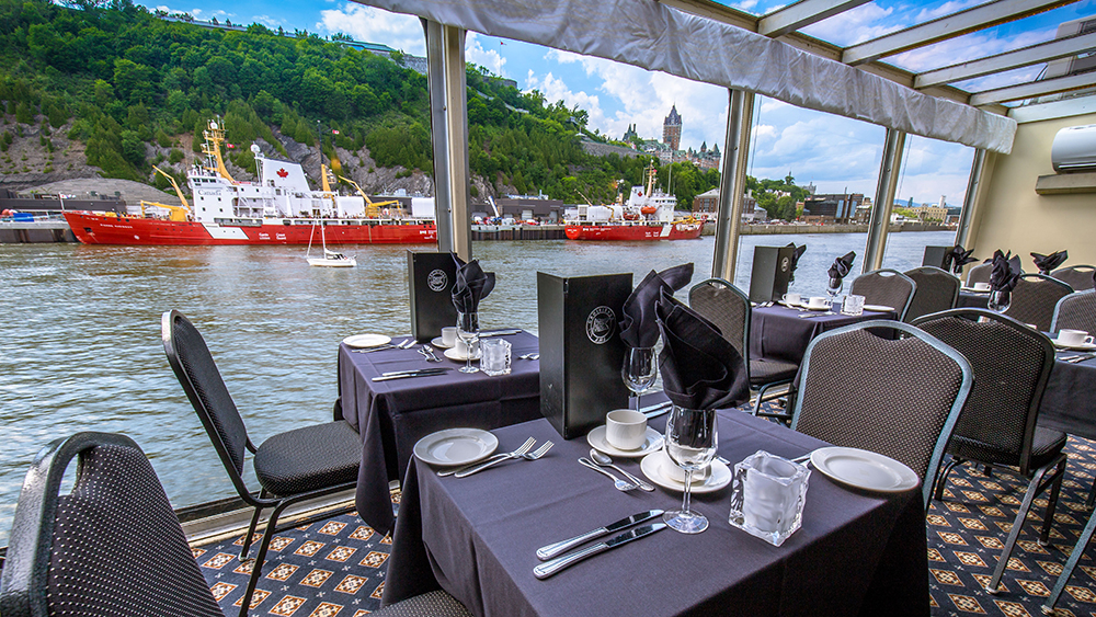 Several tables set in front of a window on the AML Louis Jolliet ship in Quebec, overlooking the river and Cap Diamant in the background