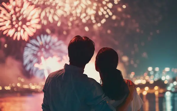 A couple watches a colorful fireworks display lighting up the night sky, with shimmering reflections on the water and city lights in the background.