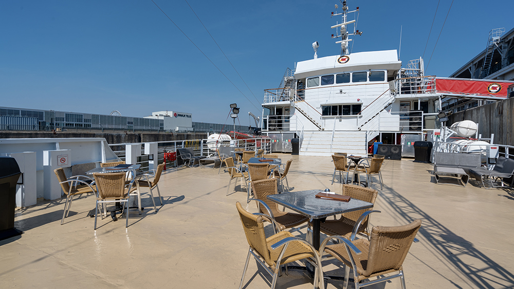 One of the outdoor terraces of the AML Cavalier Maxim ship under a blue sky. Tables and chairs are set up, and the ship is docked.