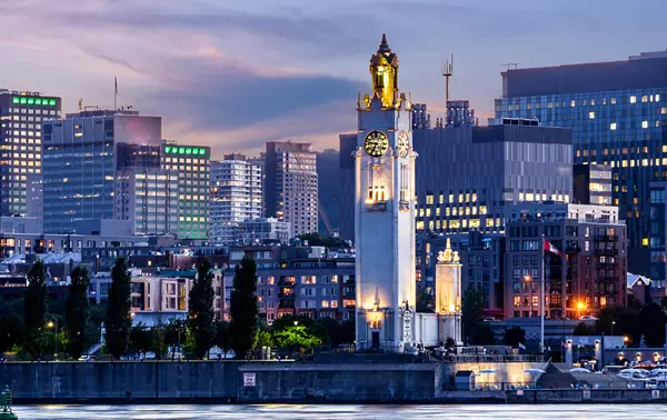The Clock Tower rises by the river, lit up at dusk, with buildings and skyscrapers illuminated in the background under a twilight sky.