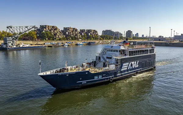 A black cruise ship with "AML" written on it, sailing on a river. In the background, a distinctive residential complex, a bridge, and urban buildings under a clear blue sky.