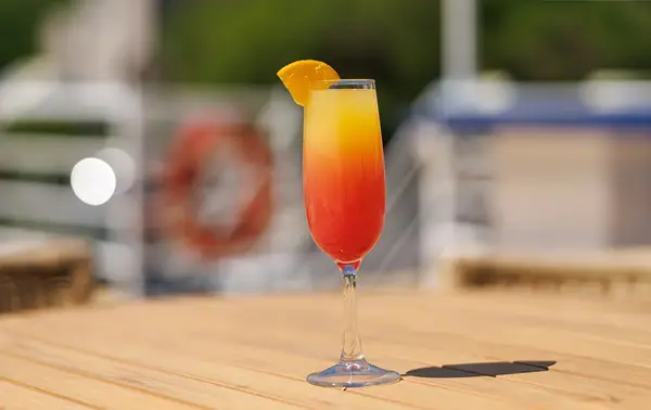 A colorful red and yellow cocktail glass garnished with an orange slice, placed on a wooden table with a blurred background featuring a boat and nautical elements.