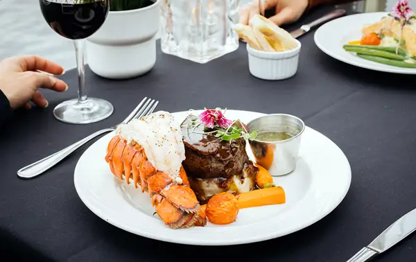 A gourmet dish featuring filet mignon, a lobster tail, grilled vegetables, and a side of sauce, served on an elegant table with a black tablecloth. A glass of red wine and another dish are also visible nearby.