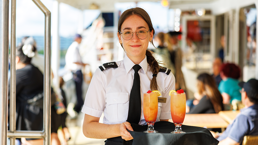 A smiling waitress in uniform holding a tray with two orange-colored cocktails on it, all aboard the AML Cavalier Maxim in Montreal.