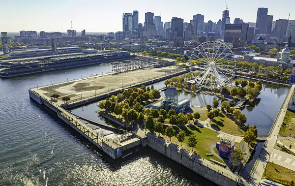Aerial view of a large urban park surrounded by water, featuring a Ferris wheel, green spaces, and modern buildings in the background of a city.