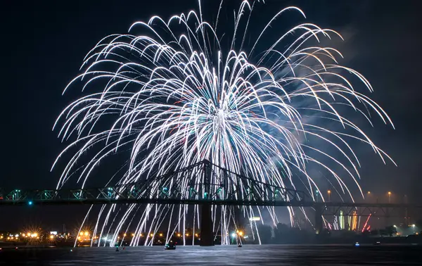 Stunning white fireworks burst in the night sky above an illuminated bridge, with shimmering reflections on the water and city lights in the background.