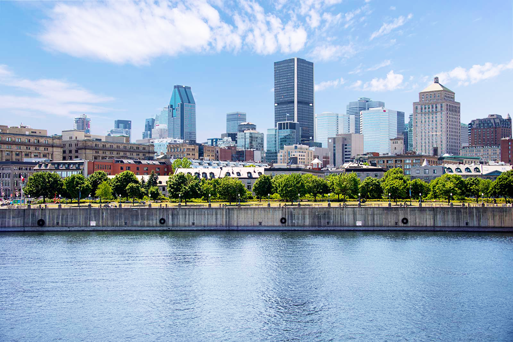 The Montreal skyline with modern buildings under a blue sky, with the St. Lawrence River in the foreground.