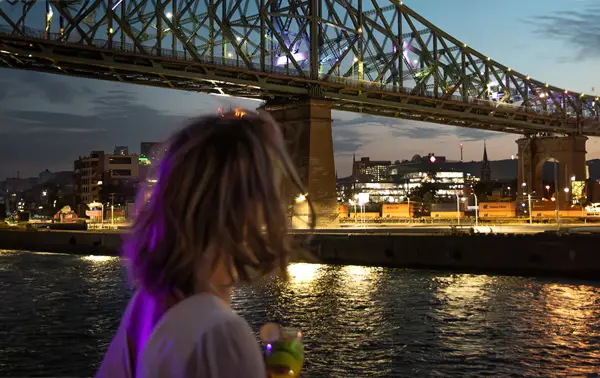 A woman viewed from behind holding a drink, admiring a lit-up bridge spanning a river, with city lights and a twilight sky in the background.
