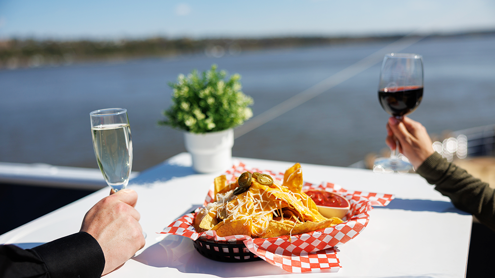 Two people sitting at a table with a view of the river, one holding a glass of wine and the other a flute of champagne, sharing a basket of nachos.