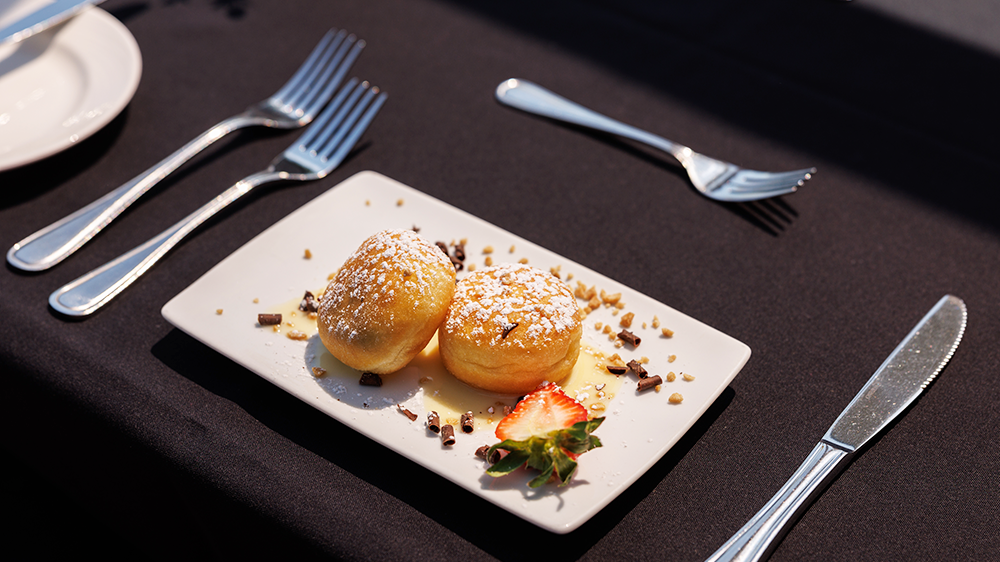 Mini beignets farcis saupoudrés de sucre glace joliment présentés dans une assiette avec une tranche de fraise fraîche et des copeaux de chocolats en décoration.