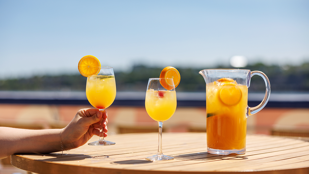 A close-up of a hand holding a glass of orange sangria, with another glass and a pitcher of sangria served on a wooden table on one of the terraces of the AML Cavalier Maxim in Montreal.