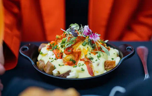 A delicious dish of roasted potatoes topped with hollandaise sauce, garnished with vegetables, fresh herbs, and edible flowers, served in a cast-iron skillet.