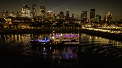 A stunning nighttime view of an illuminated cruise ship sailing on a river, with a sparkling city skyline in the background featuring skyscrapers and urban lights.