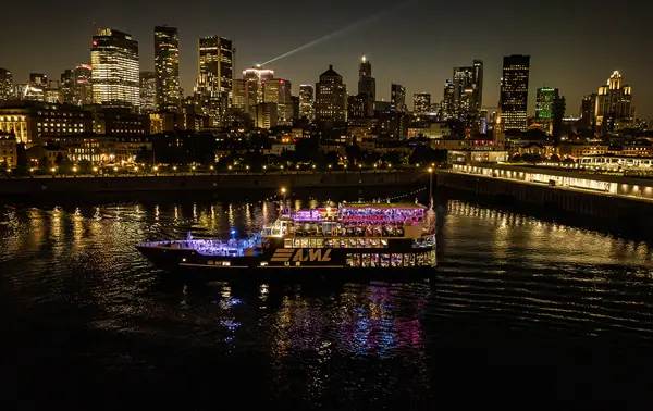 A stunning nighttime view of an illuminated cruise ship sailing on a river, with a sparkling city skyline in the background featuring skyscrapers and urban lights.