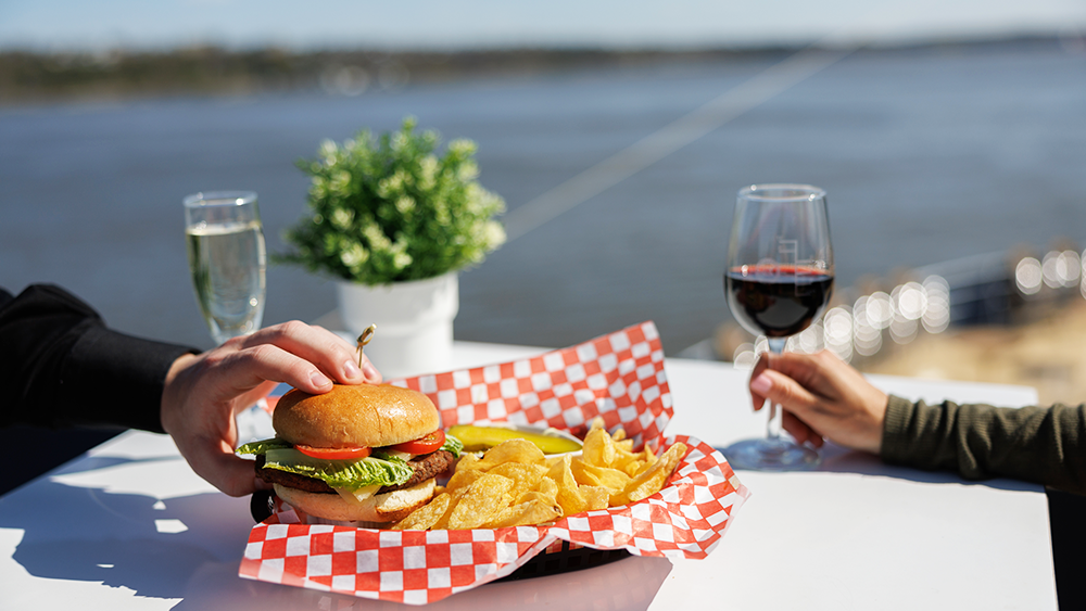 Two people enjoying a burger with fries, each with a glass of wine, on one of the terraces of the AML Cavalier Maxim in Montreal.
