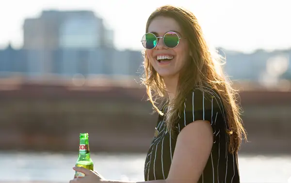 A smiling woman wearing round sunglasses and a striped shirt, holding a green beer bottle, with a river and blurred buildings in the background.