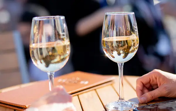 Two glasses of white wine on a wooden table on a sunny day, with visible hands and a blurred background of a lively outdoor setting.